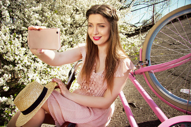 Rapariga Selfie. Retrato de sorrir jovem e bela menina de cabelos compridos em vestido rosa tomando selfie em seu telefone. Bicicleta retrô e flores no fundo
. - Foto, Imagem