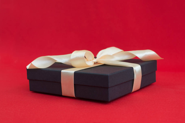 black gift box with gold ribbon and a bow on red background - Фото, зображення