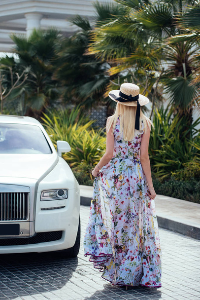 Beautiful young woman in a dress with floral pattern and a straw hat goes to the white car - Φωτογραφία, εικόνα