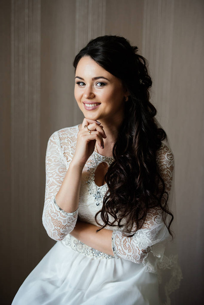 Beautiful young bride in white dress sitting and waiting for the groom indoors. Elegant charming young brunette bride is posing indoors in a wedding dress. Morning, the bride - Фото, зображення