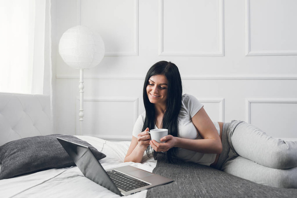 Relaxing on the bed. Beautiful woman enjoying a cup of coffee and using her laptop - Zdjęcie, obraz