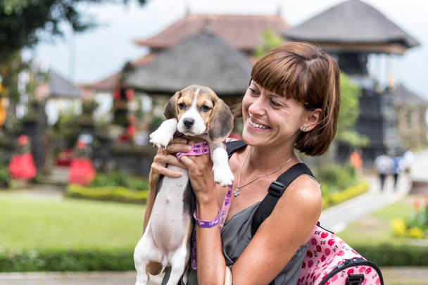 Mujer hermosa joven feliz con beagle amapola sosteniendo un perro en sus manos. El backgorund del templo balinés. Indonesia, Bali
. - Foto, imagen