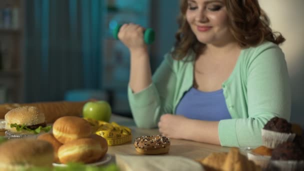 Overweight girl lifting small dumbbell and looking at donut with smirk, diet - Filmmaterial, Video