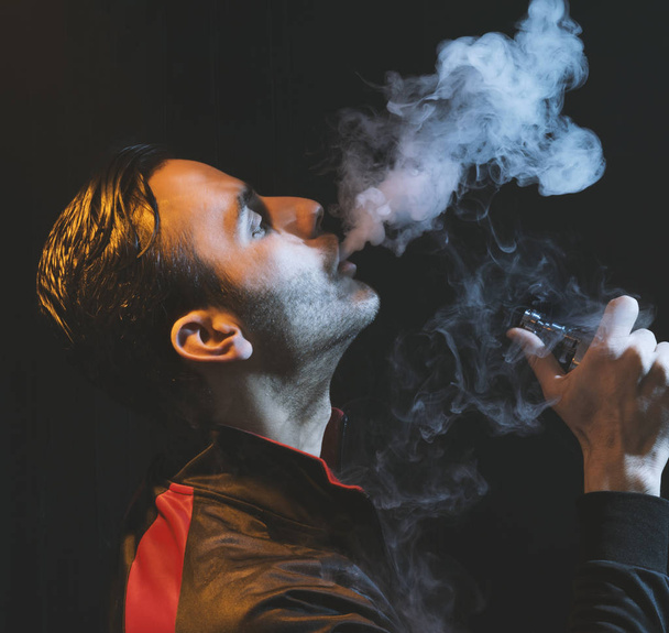 geïsoleerde man vaping e sigaret op een donkere achtergrond - Foto, afbeelding