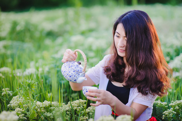 Young Chinese girl pouring tea in flowering meadow - Photo, Image