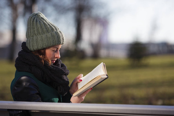 A girl in a hat sits on a park bench and reads a book. - Valokuva, kuva