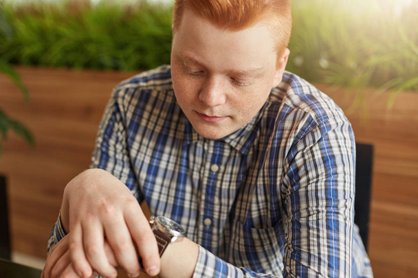 Young stylish red head man dressed in checked shirt siting in cosy cafe looking at his watch in waiting for someone. Style and fashion concept. A young businessman waiting for the meeting in cafe. - Foto, Imagem