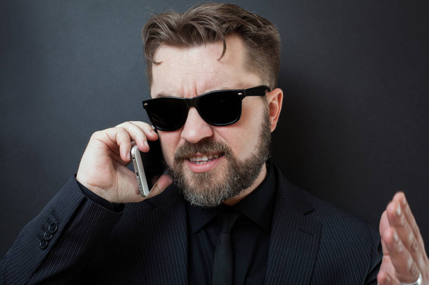 Un hombre de negocios con traje negro jura por teléfono con sus subordinados. Un hombre con gafas de sol habla por teléfono, haciendo un gesto con las manos sobre un fondo negro
 - Foto, Imagen