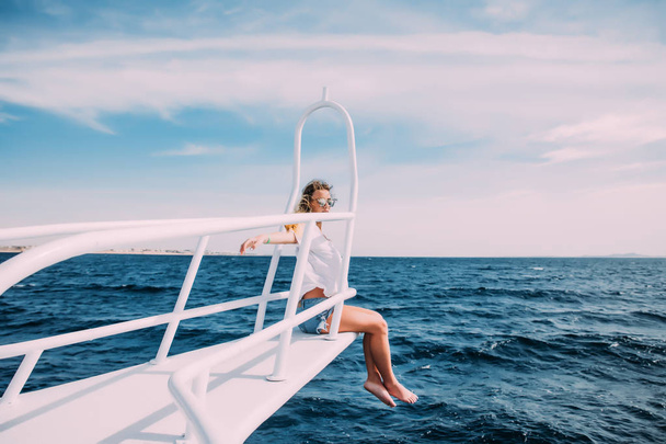 Beautiful woman in white swimsuit standing on the nose of the yacht and spreading hands apart at a sunny summer day, breeze developing hair, beautiful turquoise sea on background - Photo, Image