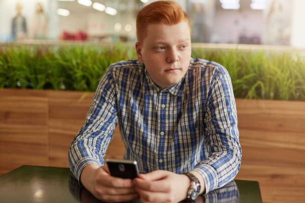 ein Porträt eines nachdenklichen Teenagers mit roten Haaren und Sommersprossen im karierten Hemd, der sein Handy am Holztisch vor grünem Hintergrund hält und geheimnisvoll zur Seite schaut. Menschen, Lebensstil - Foto, Bild