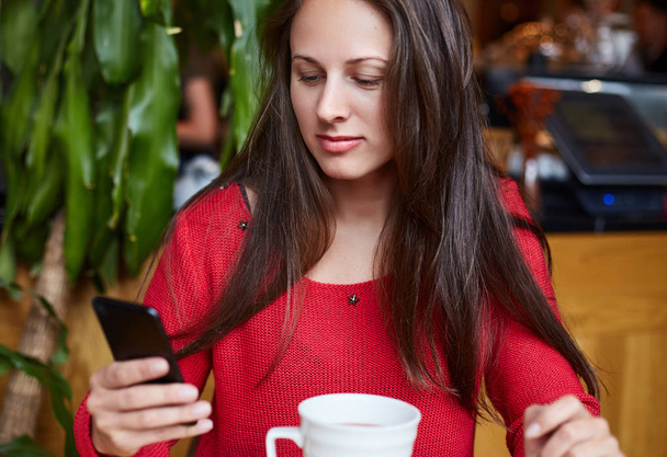 Close up shot of of beautiful young lady using her phone like a mirror or posing for selfie. Attractive woman is wearing red sweater sitting in a modern coffeehouse waiting for her lunch to come. - Photo, Image