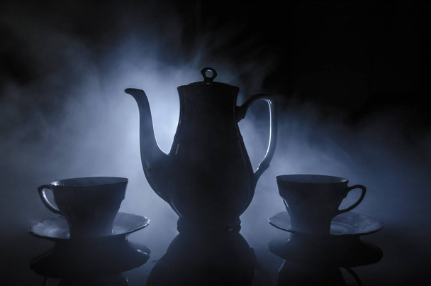 Tema del tè e del caffè. Vecchia tazza di tè o caffè in ceramica vintage con brocca e tazza di zucchero su sfondo scuro con luci e fumo. Silhouette di teiera e tazza di ceramica
. - Foto, immagini