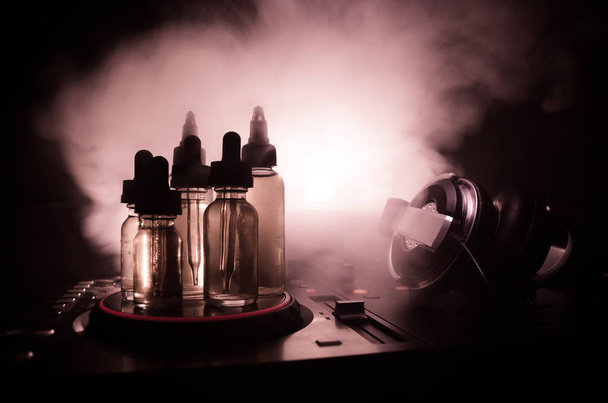 Vape and DJ Club concept. Smoke clouds and vape liquid bottles on Dj mixer close up. Light effects. Useful as background or vape or club advertisement or vape background. - Photo, Image