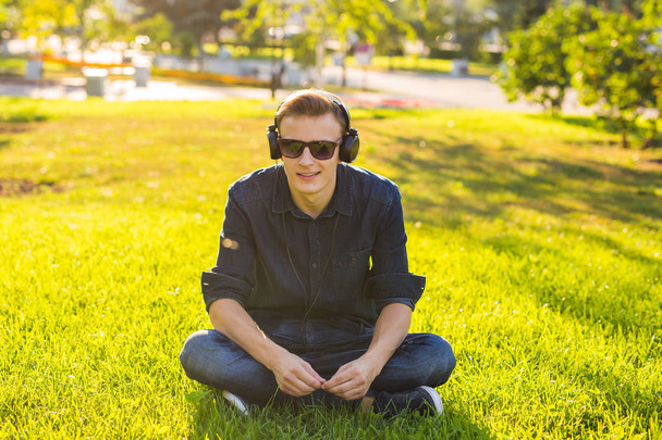 Handsome male student sitting on a green grass and listening music - Foto, Imagen