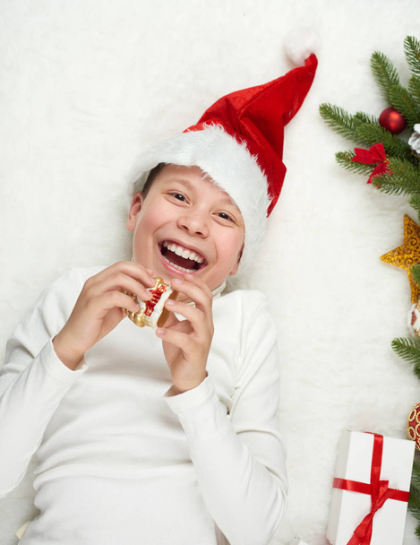 chlapec se baví s vánoční dekorace, výraz obličeje a šťastný emoce, oblečený v santa hat, leží na bílé kožešiny pozadí, Zimní dovolená concept - Fotografie, Obrázek