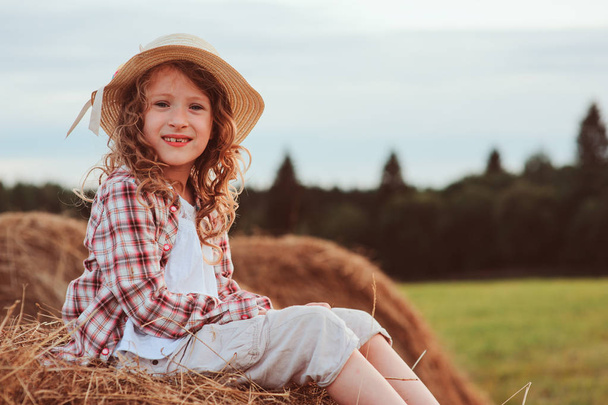 happy child girl in country style plaid shirt and hat relaxing on summer field with hay stacks - Foto, Imagem