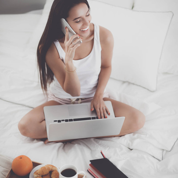 Young beautiful woman sitting in bed with laptop - 写真・画像