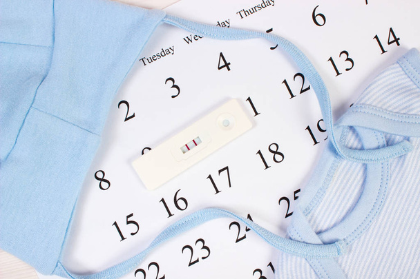 Prueba de embarazo con resultado positivo y ropa para recién nacidos en el calendario, esperando un bebé
 - Foto, imagen
