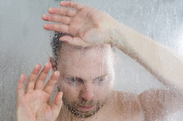 zdůraznil, muž ve sprše, stojící pod tekoucí vodou a klade ruku na sklo ve sprše v kabině za dveřmi transparentní zapocené sklo v koupelně - Fotografie, Obrázek