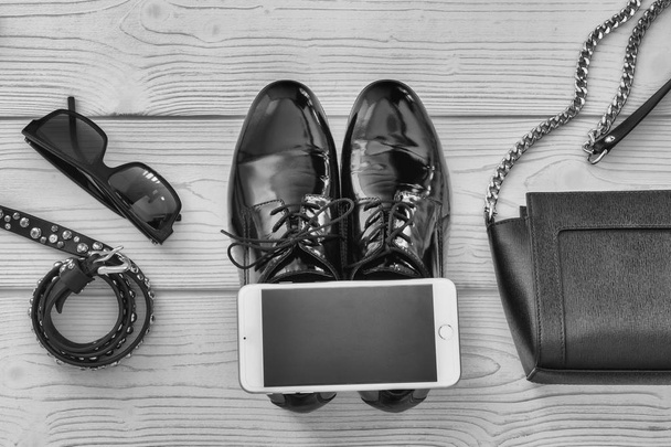 Elegantes Set aus Gürtel, Brille, Tasche, Telefon und Businessschuhen von oben. Bild von modischen Schuhen und Accessoires mit einem hölzernen Hintergrund für Text. Schwarz-Weiß-Image von Modeaccessoires. - Foto, Bild