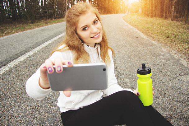 Selfie time. Portrait of pretty and young redhaired sports girl taking a selfie while sitting on the park road. - Valokuva, kuva