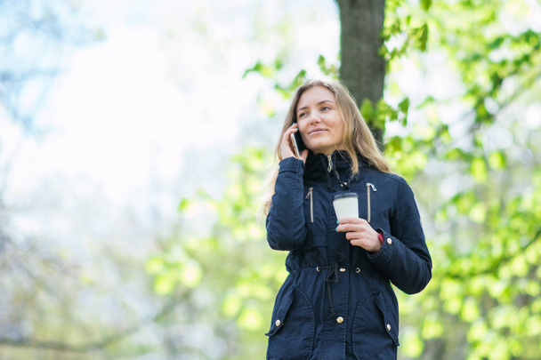 Μιλάτε στο τηλέφωνο, ένα κορίτσι είναι το περπάτημα το πρωί σε ένα πάρκο με ένα smartphone και καφέ - Φωτογραφία, εικόνα