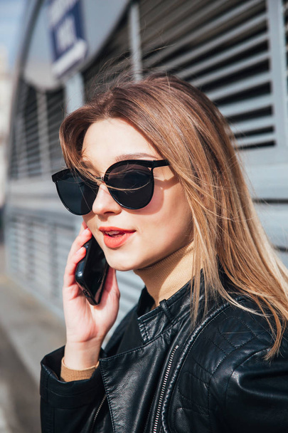Fashion portrait pretty woman in black rock style in sunglasses using mobile phone over gray background in city - Foto, imagen