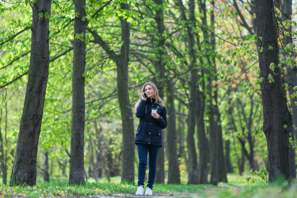 Praten over de telefoon, loopt een meisje in de ochtend in een park met een smartphone en koffie - Foto, afbeelding