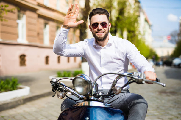 Веселый молодой бородатый мужчина в шлеме сидит на скутере и здоровается на городской улице
 - Фото, изображение