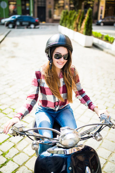 Szczęśliwa młoda kobieta jeździć na skuterze w okularach słonecznych i hemlet na zewnątrz patrząc na kamery - Zdjęcie, obraz
