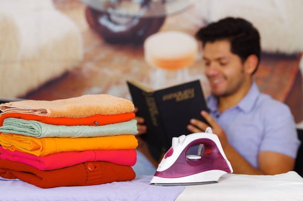 A pile of clothes next to the iron with gorgeous man smiling behind while he is reading a book - 写真・画像