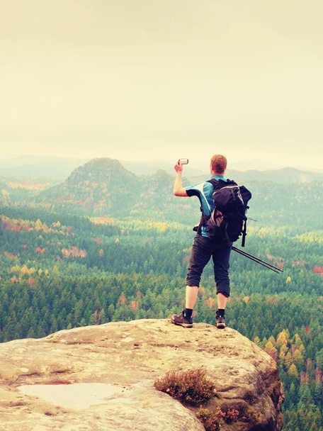 Імбирний чоловік робить фотографії зі смартфоном на скелястому піку над автономною долиною
. - Фото, зображення