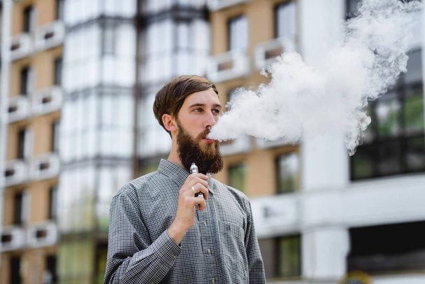 Men model with beard vaping an electronic cigarette. Vaping outdoors. Safe smoking. Young hipster vaper. - Photo, Image
