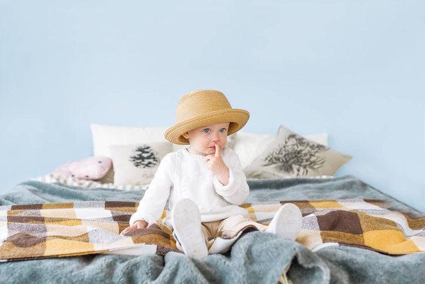 Dość mały dziewczyna w słomkowym kapeluszu, z niebieskimi oczami i wyrażenie zadumany, siedząc na łóżku - Zdjęcie, obraz