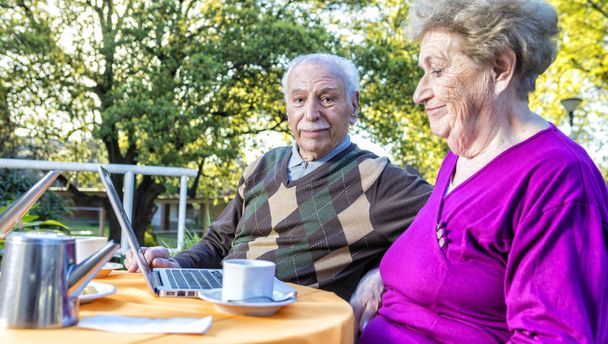 Iloinen vanhempi pari tekee aamiaista ulkona ja kannettavan tietokoneen avulla
 - Valokuva, kuva