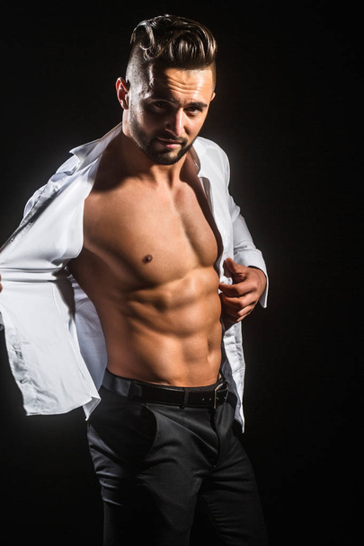 Attractive sports man takes off his shirt. Businessman with beautiful strong body. Passion lover. Young man with naked torso on black background looks into the camera. Clothes for business people - Фото, изображение