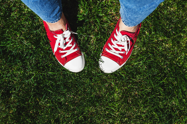 Τα πόδια στην παλιά κόκκινα αθλητικά παπούτσια για την πράσινη χλόη. Θέα από ψηλά. Το co - Φωτογραφία, εικόνα