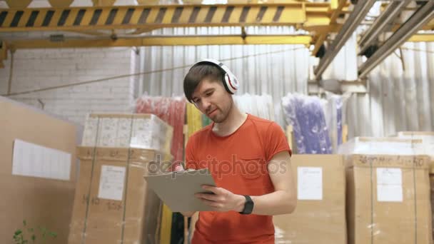Dolly shot of Happy young worker in industrial warehouse listening to music and dancing during work. Man in headphones have fun at workplace. - Footage, Video