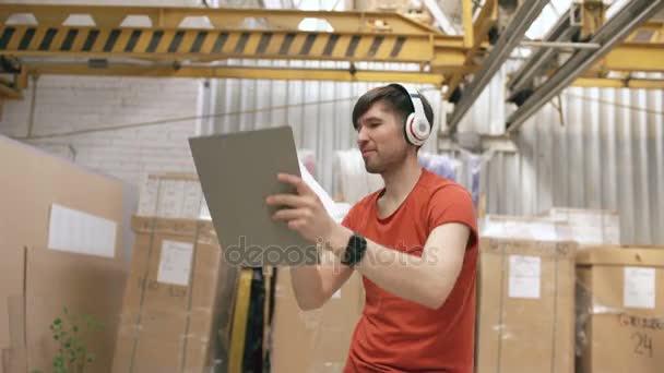 Dolly schot van Happy jonge werknemers in industriële magazijn luisteren naar muziek en dansen tijdens werk. Man in hoofdtelefoons hebben plezier op werkplek. - Video