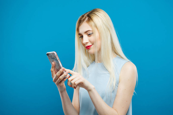 Die erstaunliche Blondine mit langen Haaren und sinnlichem Blick berührt den Bildschirm ihres Smartphones auf blauem Hintergrund. Nettes Mädchen mit Handy im Studio. - Foto, Bild