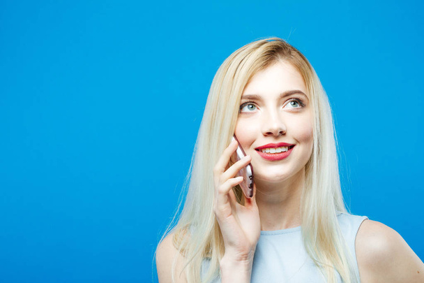 Portrait of Smiling Blonde with Sensual Lips Talking with Somebody by Her Smartphone on Blue Background. Cute Girl with Mobile in Studio. - Foto, Bild