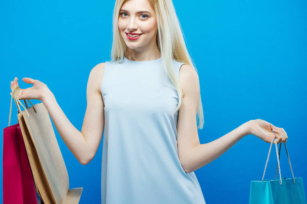 Happy Blonde with Lond Hair and Charming Smile on Blue Background in Studio. Satisfied Woman Holding a Lot of Shopping Colorful Bags. - Foto, afbeelding