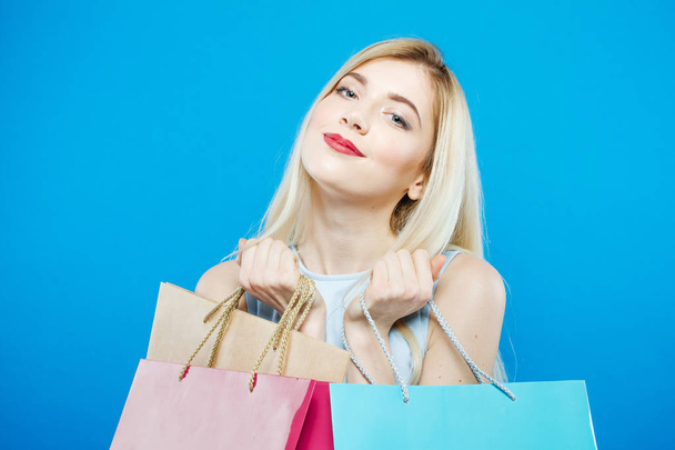 Smiling Satisfied Woman Holding a Lot of Shopping Colorful Bags in Studio. Happy Blonde with Lond Hair and Charming Smile on Blue Background. - Foto, Imagem