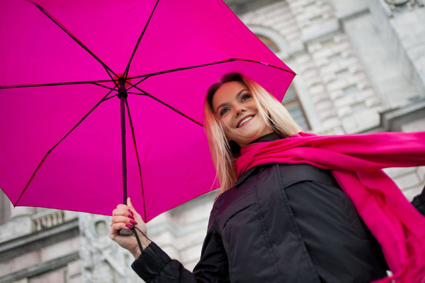 Beautiful young and happy blond woman with colorful umbrella on the street. The concept of positivity and optimism. Girl in a bright pink scarf and umbrella walking in a rainy city. - Foto, Imagem