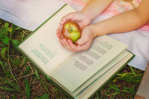 Τα χέρια των παιδιών που κατέχουν ένα μήλο σε ένα ανοικτό βιβλίο στη φύση. - Φωτογραφία, εικόνα