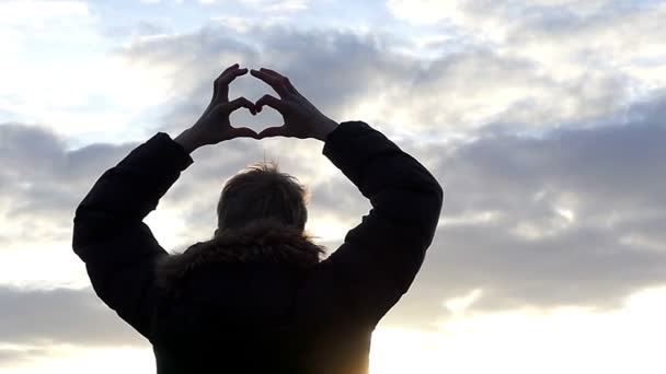 Σιλουέτα ενός αγοριού με την πλάτη στην κάμερα κρατά τα χέρια σε σχήμα καρδιάς ενάντια στον όμορφο ουρανό σε αργή κίνηση - Πλάνα, βίντεο