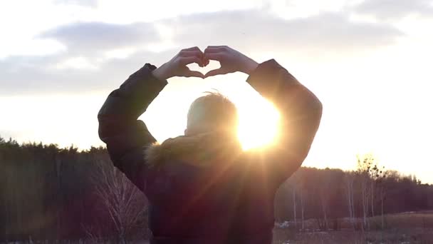 το αγόρι με την πλάτη στην κάμερα κρατά τα χέρια του στο σχήμα της καρδιάς για το Backgroung του ηλιόλουστου ουρανού σε αργή κίνηση - Πλάνα, βίντεο