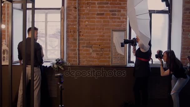 Veel fotografen uit de masterclass nemen een foto van een mooie paar modellen in een zwarte trouwjurk en bugs op de achtergrond van een rode bakstenen wal. - Video