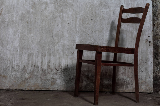 Old worn chair with cracks is on a concrete floor in an abandoned, gloomy room with a dim light - Photo, Image
