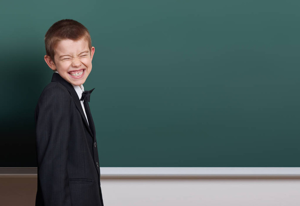 elementary school boy make faces near blank chalkboard background, dressed in classic black suit, one pupil, education concept - Zdjęcie, obraz